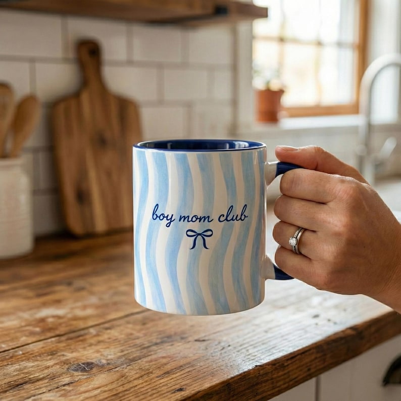 May include: A white ceramic mug with blue wavy stripes and a blue interior. The mug has the text "boy mom club" in blue script with a bow graphic. The mug is held by a person.