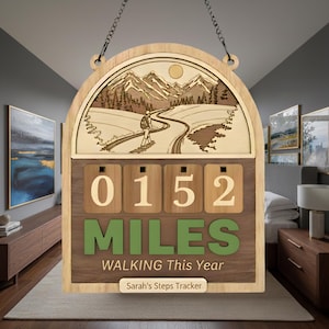 Puede incluir: Contador de pasos de pared de madera con una escena de montaña y un excursionista. El contador muestra "0152 Miles Walking This Year". El rastreador está personalizado con "Sarah's Steps Tracker" en la parte inferior. Cuelga de una cadena.