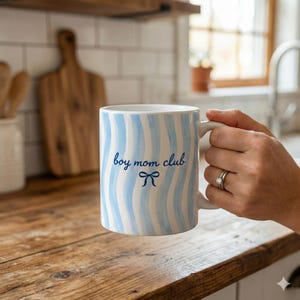 May include: White ceramic mug with blue wavy stripes and the text "boy mom club" in a script font, along with a small bow graphic. The mug is held by a person's hand, set against a kitchen backdrop.
