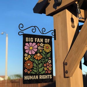 May include: A rectangular black garden flag hanging from a black metal scroll bracket. The flag features the text "BIG FAN OF HUMAN RIGHTS" in a bold, light-colored font, surrounding a central illustration of various colorful flowers.
