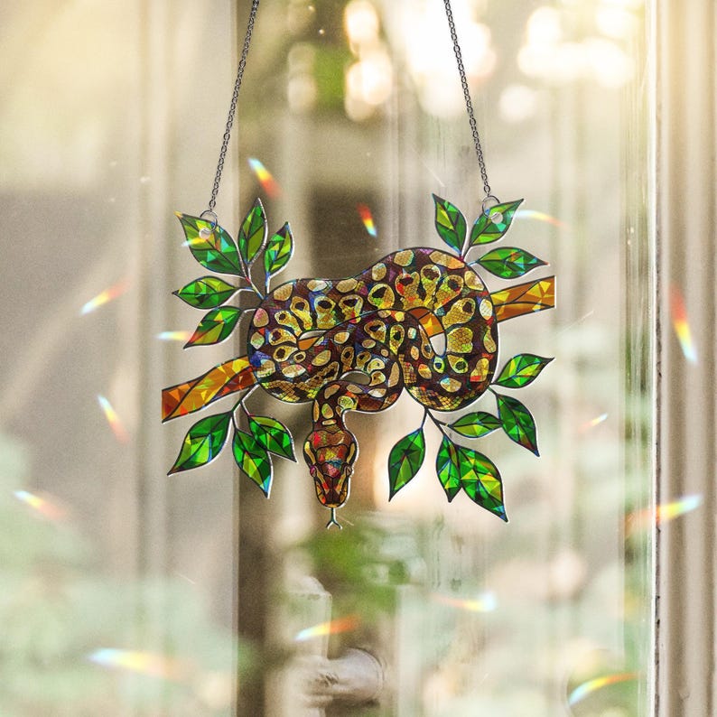 May include: Stained glass window hanging depicting a snake coiled around a branch with green leaves. The snake has a brown and yellow pattern. The piece is suspended by a chain.