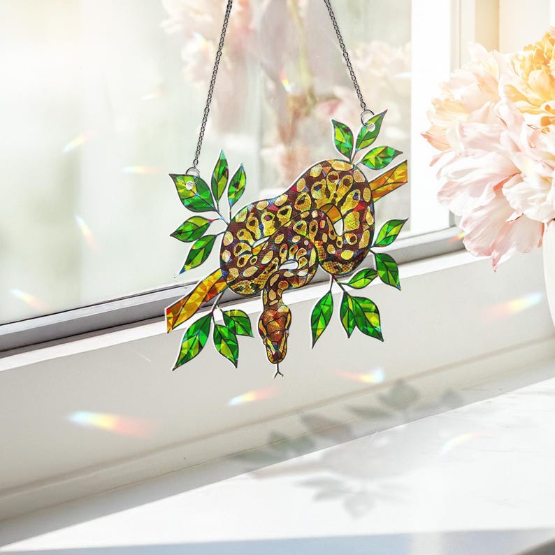 May include: A stained glass snake suncatcher, featuring a brown, yellow, and red patterned snake coiled around a yellow branch with green leaves. It hangs from a silver chain, suitable for window or wall decor. A unique piece of art.