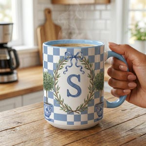 May include: A large ceramic mug with a blue and white checkered pattern. The mug features a blue letter "S" inside a wreath of olive branches and a blue ribbon. Two potted topiary trees are on the sides. The handle and rim are light blue.