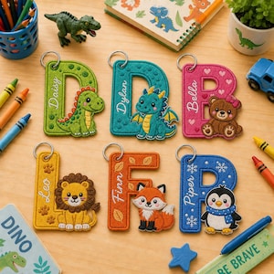 Op de afbeelding: Zes geborduurde letter-sleutelhangers met cartoon-dieren en namen. De letter D heeft een groene dinosaurus, D een groenblauwe draak, B een bruine beer, L een gele leeuw, F een oranje vos en P een blauwe pinguïn met sneeuwvlokken.