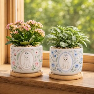 May include: Two cylindrical ceramic planters on wooden saucers. The left planter features pink flowers and a white cat illustration labeled Kiki. The right planter holds a green leafy plant and features blue flowers and a white cat labeled Kiki.