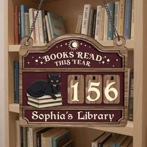 May include: Wooden sign with the text "BOOKS READ THIS YEAR" and "Sophia's Library". A black cat is on a stack of books. The counter reads 156. The sign is hanging in front of a bookshelf.