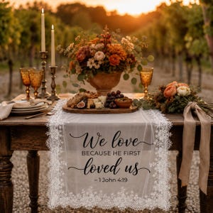 以下が含まれることがあります： 黒い文字で「We love BECAUSE HE FIRST loved us - 1 John 4:19」とプリントされた白いレースのテーブルランナー。木製のテーブルの上に敷かれ、花のアレンジメント、金属製のホルダーに入った2本の背の高いキャンドル、グラスが置かれています。