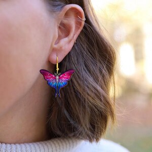 May include: A pair of colorful moth earrings with gold-colored hooks. The moth design features a gradient of pink, purple, and blue hues. The earrings are a unique accessory.