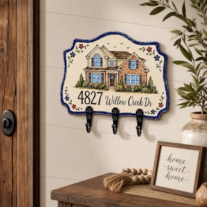 May include: A rectangular wall-mounted key holder with a scalloped blue border and floral accents. It features a painted illustration of a two-story brick house and the text 4827 Willow Creek Dr. Three black metal hooks are attached to the bottom.
