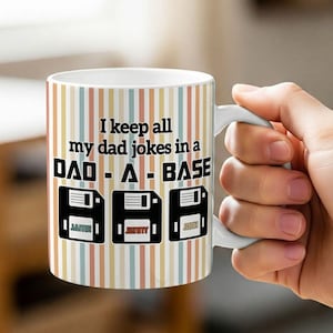 May include: White ceramic mug with a pastel striped design. The mug has the text "I keep all my dad jokes in a DAD - A - BASE" with floppy disk graphics and names. The mug is held by a hand.