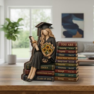 May include: A figurine of a graduate wearing a black cap and gown, holding a diploma and a sunflower bouquet. The figure sits on a stack of books labelled Emma, Custom School Name, and Class of 2026, next to a stack of books with inspirational text.