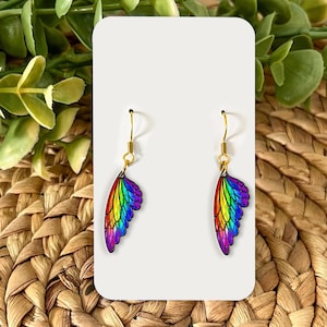 May include: A pair of earrings with a wing design in a rainbow colour scheme. The wings are detailed with shades of red, orange, yellow, green, blue, and purple. The earrings have gold-coloured hooks.