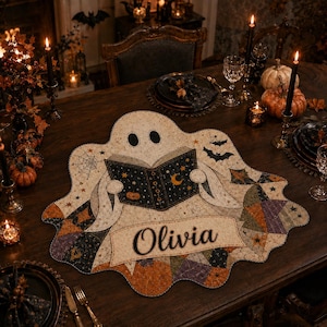 Peut inclure: Un set de table en tissu en forme de fantôme avec un motif imprimé représentant un fantôme lisant un livre orné de motifs célestes, dont des étoiles, un croissant de lune et une toile d'araignée. Le prénom Olivia est inscrit en noir.