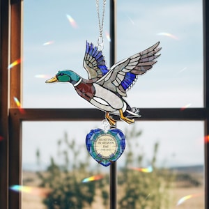 May include: A stained glass-style duck suncatcher with outstretched wings, featuring a green head, brown chest, and blue and gray wings. A heart-shaped pendant below reads "Hunting in Heaven Dad 1938-2023". The suncatcher hangs from a silver chain.
