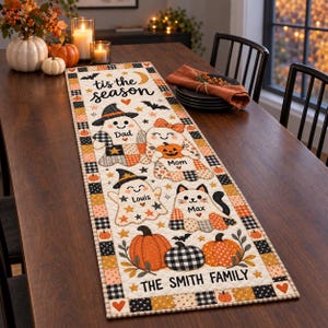 Op de afbeelding: Een rechthoekige gewatteerde tafelloper met een crèmekleurige achtergrond en een herhalende rand van oranje, zwarte en witte vierkanten. Het ontwerp bevat spookfiguren met de labels Dad, Mom, Louis en Max, pompoenen, vleermuizen en de tekst 'tis the season' en 'THE SMITH FAMILY'.