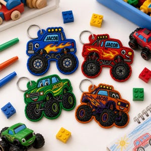 May include: Four embroidered monster truck keychains in blue, red, green, and orange, each featuring the name JACOB in black text, large black tyres, and a silver metal keyring attached to the top of the truck cab for hanging or attachment.