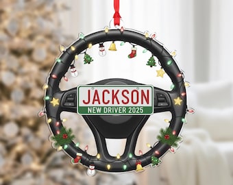 Pode incluir: Um enfeite de Natal em forma de volante preto, adornado com luzes festivas e pequenas decorações de férias. O enfeite apresenta uma placa de matrícula com o nome "Jackson" e "NEW DRIVER 2025". Uma fita vermelha é anexada para pendurar.
