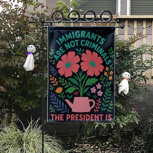 Op de afbeelding: Een zwarte tuinvaandel met de tekst "IMMIGRANTS ARE NOT CRIMINALS" en "THE PRESIDENT IS". De vlag heeft een bloemenontwerp met twee grote roze bloemen, een roze gieter en diverse andere bloemen en bladeren.