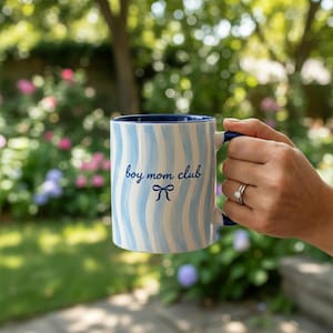 May include: A white ceramic mug with blue wavy stripes and a blue interior. The mug has the text "boy mom club" and a bow graphic in blue. The mug is held in front of a blurred garden background.