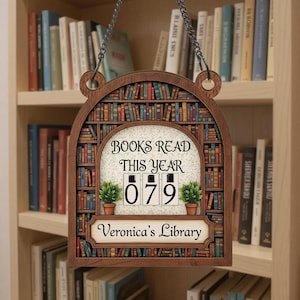 Puede incluir: Un letrero de madera con diseño de estantería, que muestra "BOOKS READ THIS YEAR" y el número "079". El letrero incluye el nombre "Veronica's Library" y dos pequeñas plantas en macetas. Cuelga de una cadena de metal.