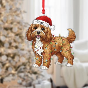 May include: A brown and white dog ornament with a red Santa hat that says "Max". The dog is decorated with a string of colorful Christmas lights and a red ribbon for hanging. The ornament is a festive holiday decoration.