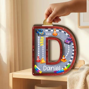 May include: A wooden letter D shaped piggy bank with a dark wood frame and a clear front panel. The interior features a race track graphic with a blue race car, a purple race car, orange traffic cones, and the name Daniel printed in white text.