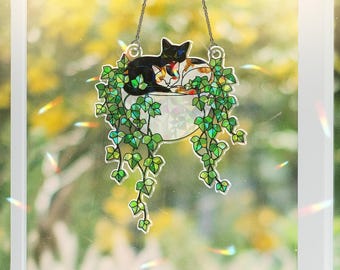 Puede incluir: Colgante de ventana de vidriera con un gato negro y otros dos gatos durmiendo en una maceta blanca con hiedra verde. El arte de la ventana está suspendido por una cadena plateada, con un fondo borroso de una escena exterior soleada.