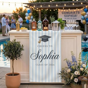 May include: A light blue and white striped table runner featuring a graduation cap icon and the text "CHEERS TO Sophia CLASS OF 2026". The runner is displayed on a white bar counter with bottles, a metal bucket, and wine glasses.