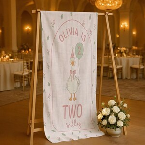 May include: A linen banner on a wooden easel displays a cartoon goose holding a balloon. The banner reads "Olivia is Two Silly" in pink script. The goose wears a party hat and bow. Floral accents and a pink border complete the design.