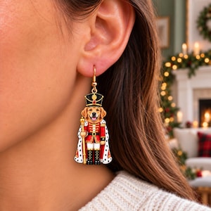 May include: A dangling earring featuring a golden retriever dog dressed as a nutcracker. The dog wears a red military-style jacket with gold trim, a black hat with a red gem, and a white cape with black spots, while holding a gold staff.