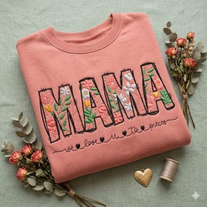 Peut inclure: Un sweat-shirt corail avec le mot "MAMA" brodé en grandes lettres fleuries. La phrase "we love u to pieces" est brodée en dessous. Le design comprend diverses fleurs et feuilles. Le sweat-shirt est de style col rond.
