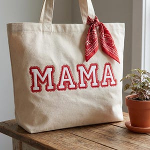 Peut inclure: Un sac fourre-tout en toile beige avec le mot "MAMA" brodé en rouge et blanc. Un bandana rouge est noué à l'une des poignées. Le sac est posé sur une surface en bois.