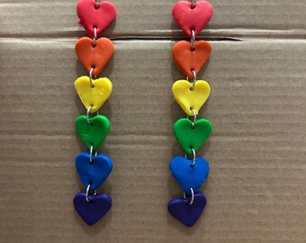 Könnte beinhalten: Regenbogenförmige Ohrringe mit Herzanhängern. Jeder Ohrring hat sechs Herzen in den Farben Rot, Orange, Gelb, Grün, Blau und Lila.
