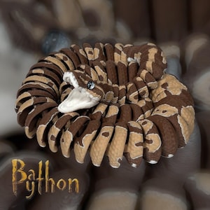 May include: A coiled plush ball python in shades of brown and tan, with a white mouth and a single, sparkling blue eye. The snake is designed with segmented body sections. The text "Ball Python" is written in a stylized font.