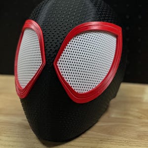 May include: A black full-head mask featuring a textured, carbon-fiber-like surface pattern. The mask includes two large, angular eye openings outlined in bright red plastic frames, with white mesh material covering the eye lenses for visibility.