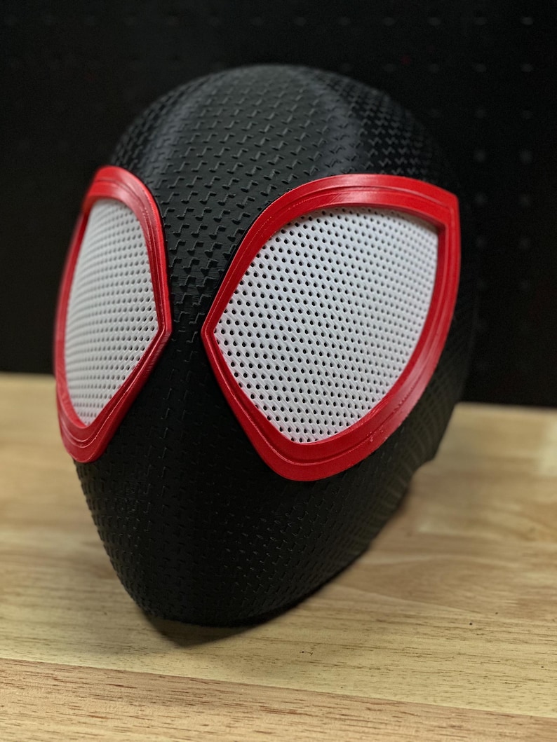 May include: A black full-head mask featuring a textured, carbon-fiber-like surface pattern. The mask includes two large, angular eye openings outlined in bright red plastic frames, with white mesh material covering the eye lenses for visibility.