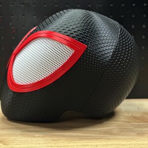 May include: A black helmet featuring a textured, dimpled surface pattern across the exterior. The helmet includes a prominent eye-shaped opening outlined in a bold red frame, containing a white, perforated mesh screen covering the eye area.
