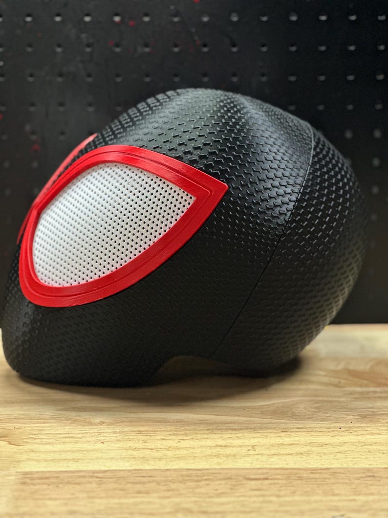 May include: A black helmet featuring a textured, dimpled surface pattern across the exterior. The helmet includes a prominent eye-shaped opening outlined in a bold red frame, containing a white, perforated mesh screen covering the eye area.