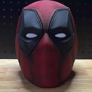 Könnte beinhalten: Eine rot-schwarze Deadpool-Maske. Die Maske hat eine strukturierte Oberfläche und verfügt über große schwarze Augenabdeckungen mit weißen Netzkopföffnungen. Die Maske ist eine vollständige Kopfbedeckung.