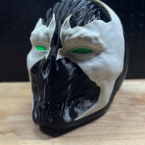 May include: A three-dimensional mask featuring a high-contrast black and white design. The mask has a glossy black base with white facial overlays and bright green mesh eye coverings. The surface displays visible layered textures from production.