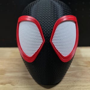 May include: A black full-head mask featuring a textured surface pattern and two large, almond-shaped eye openings. Each eye opening is outlined with a bold red frame and filled with a white, fine-mesh material for visibility.
