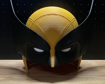 Könnte beinhalten: Eine gelb-schwarze Superheldenmaske mit einer texturierten Kuppel, zwei hohen, spitzen Seitenflossen und weißen Augenausschnitten. Die Maske hat eine glatte, matte Oberfläche an den schwarzen Teilen und eine gitterartige Textur am gelben Oberteil.