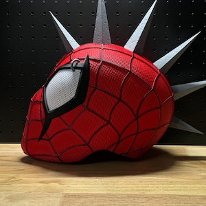 May include: A red and black Spider-Man mask with a web pattern, featuring a white mesh eye covering and gray spikes. The mask is set against a black background and rests on a wooden surface.