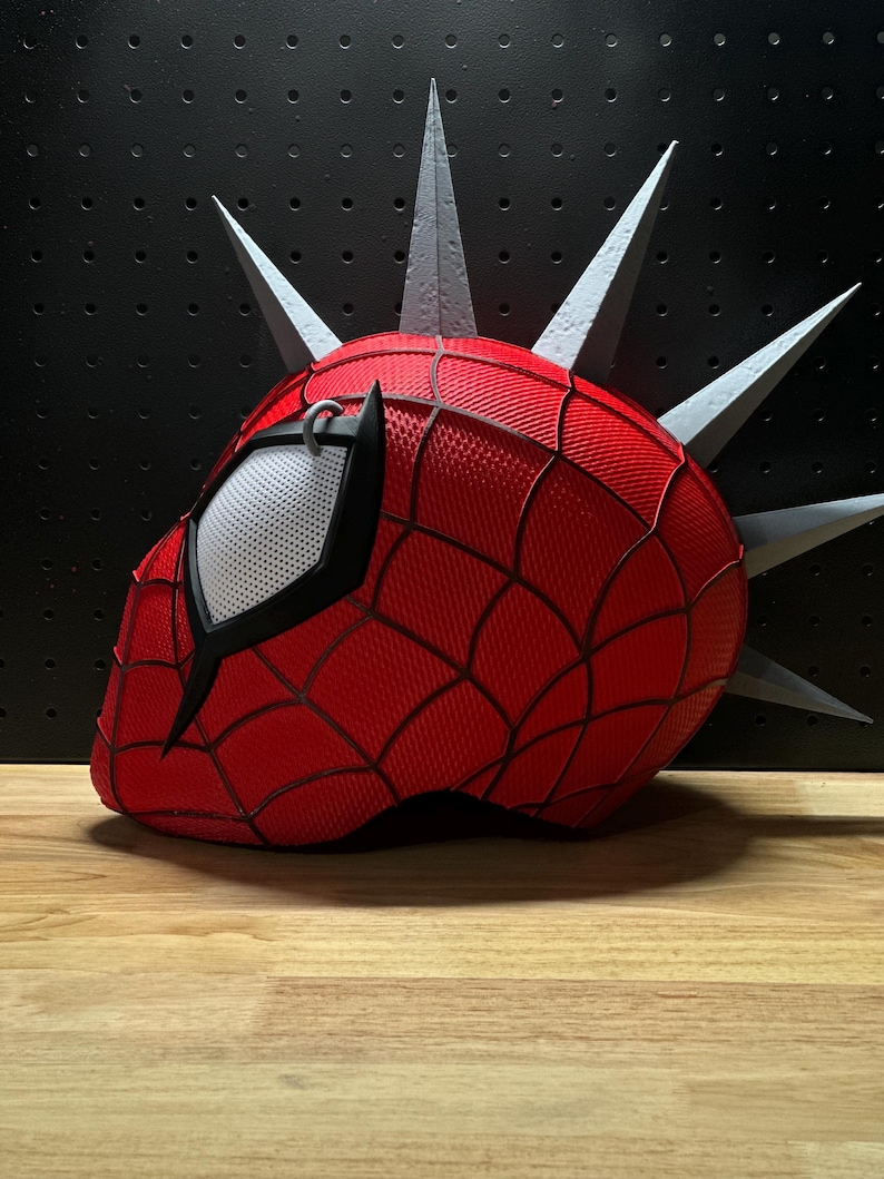 May include: A red and black Spider-Man mask with a web pattern, featuring a white mesh eye covering and gray spikes. The mask is set against a black background and rests on a wooden surface.