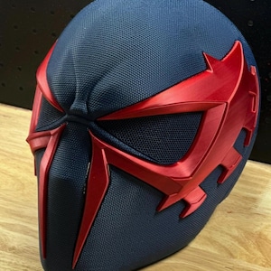 May include: A navy blue and red superhero mask with a textured surface. The mask features red accents around the eyes and face, with a mesh eye covering. The design is angular and futuristic, suggesting a comic book or cosplay theme.
