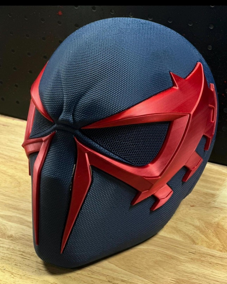 May include: A navy blue and red superhero mask with a textured surface. The mask features red accents around the eyes and face, with a mesh eye covering. The design is angular and futuristic, suggesting a comic book or cosplay theme.