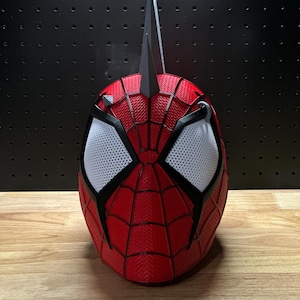 May include: A red and black Spider-Man mask with white mesh eye coverings. The mask has a textured surface and a web-like pattern. Two black, pointed structures extend from the top of the mask.