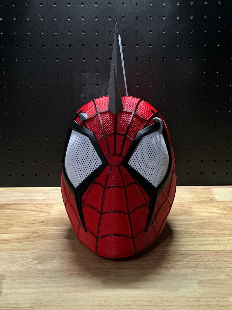 May include: A red and black Spider-Man mask with white mesh eye coverings. The mask has a textured surface and a web-like pattern. Two black, pointed structures extend from the top of the mask.