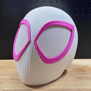 Könnte beinhalten: Eine weiße Vollkopfmaske mit einer texturierten, gitterartigen Oberfläche und zwei großen, eckigen Augenöffnungen, die mit leuchtendem Pink umrandet sind. Die Maske hat eine glatte, abgerundete Form und bedeckt den gesamten Gesichtsbereich.