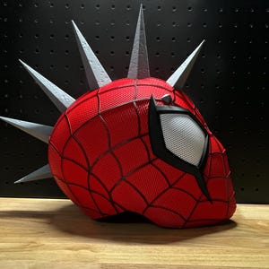 May include: A red Spider-Man mask with a web pattern and a white eye. The mask has silver spikes around the head. The mask is on a black surface.